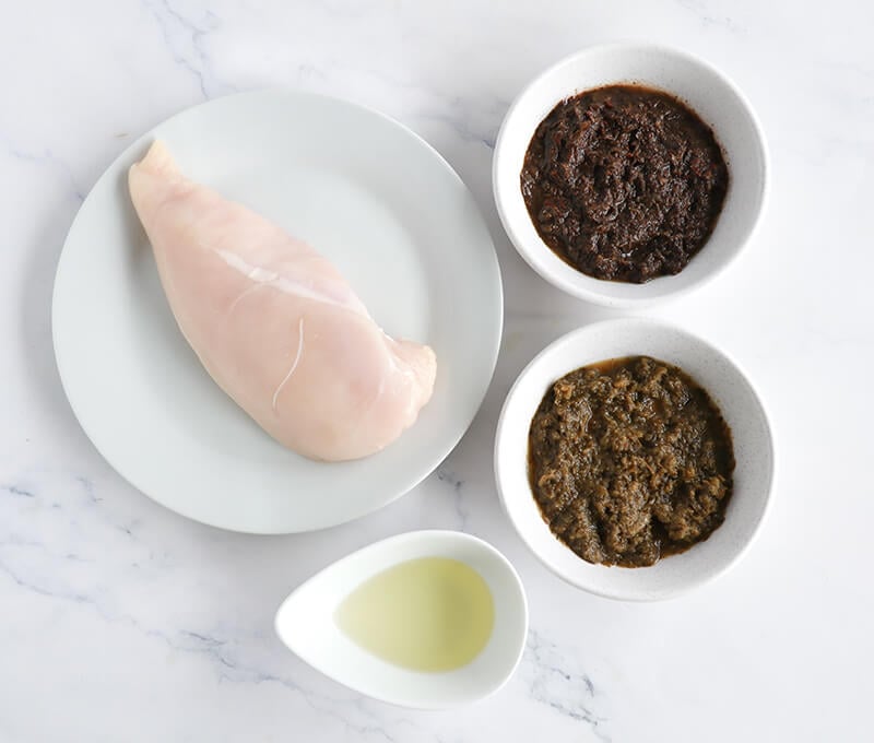 Flatlay of ingredients for jerk chicken including raw chicken breast, green seasoning, jerk marinade, and oil.