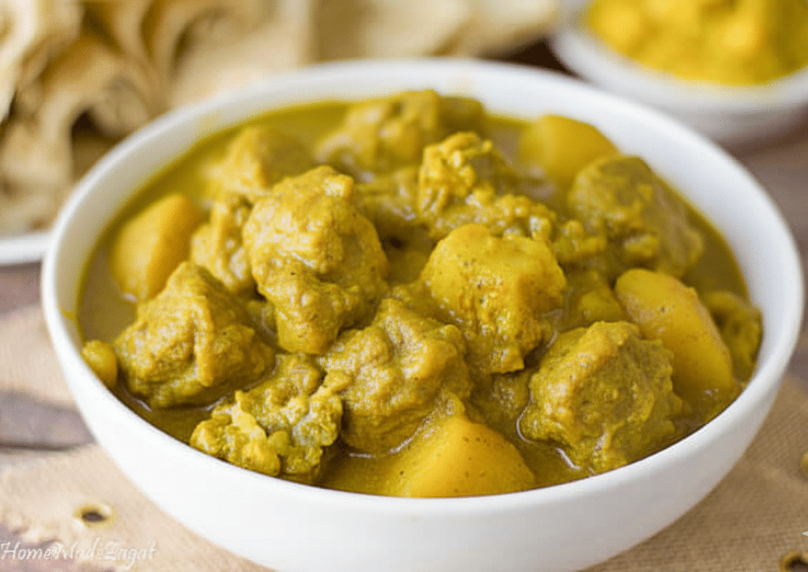 A delicious curry chicken dish, infused with various spices that can be eaten with rice or with roti. Tender chunks of curry chicken and potatoes simmered in golden Caribbean curry sauce, served in a white bowl with roti in the background.