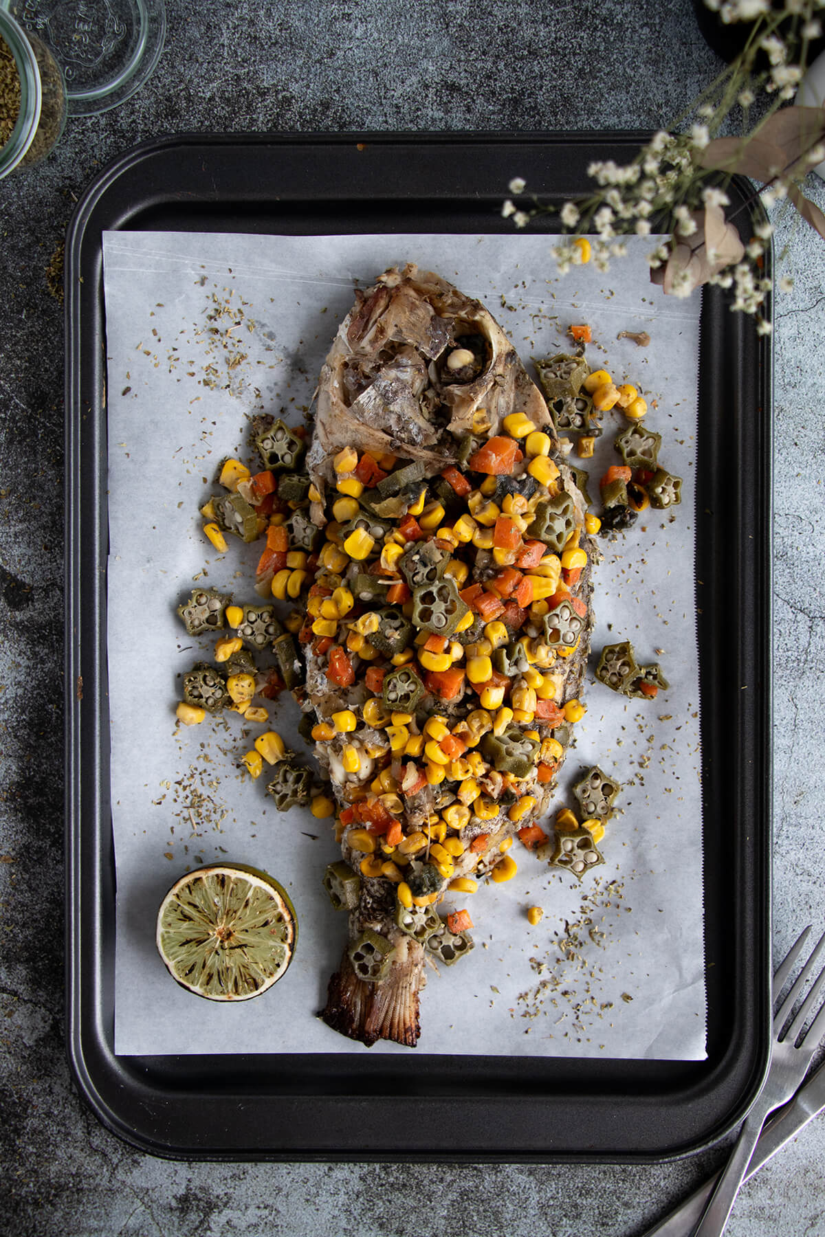 Caribbean-style whole stuffed snapper topped with okra, corn, and carrots on a baking tray lined with parchment paper.