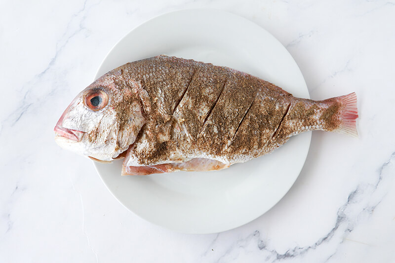 Whole snapper seasoned with salt and black pepper on a white plate, ready to be stuffed and baked Caribbean style.