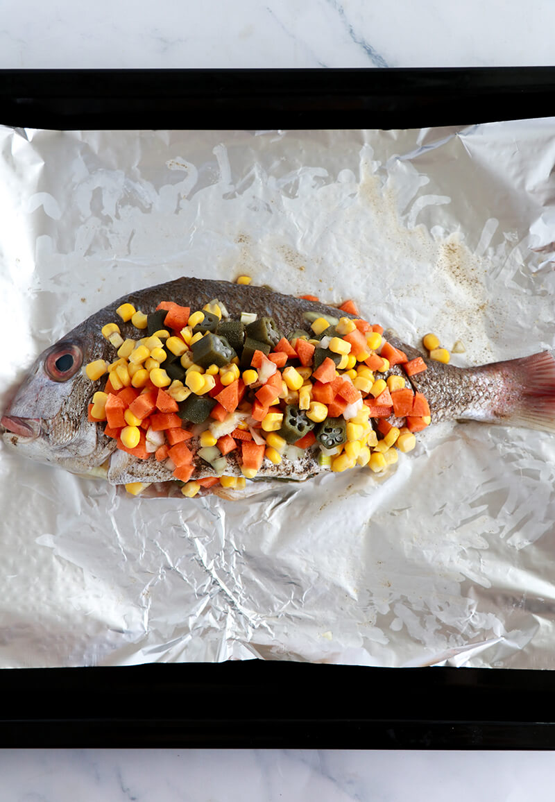 Whole fish stuffed with okra, corn, and carrots on foil, ready to be wrapped and oven-baked Caribbean style.