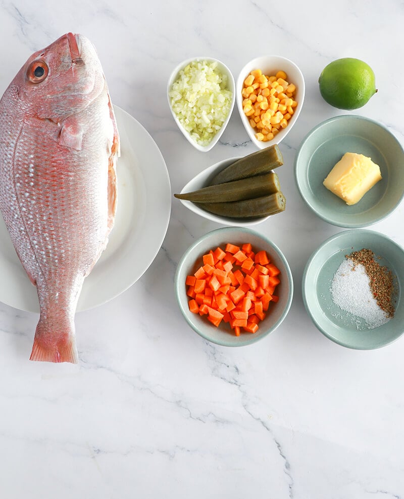 Ingredients for Caribbean-style whole stuffed fish including fresh snapper, lime, butter, okra, sweet corn, onion, carrot, salt, and pepper.
