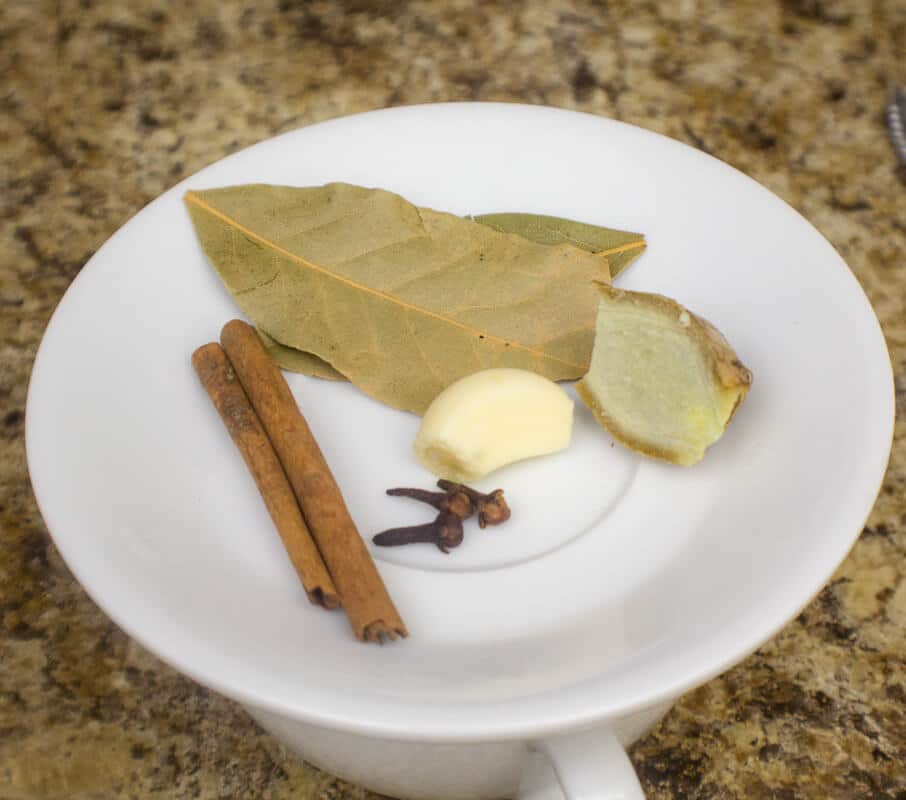 Ingredients for Caribbean bush tea including bay leaves, ginger, garlic, cloves, and cinnamon on a white mug