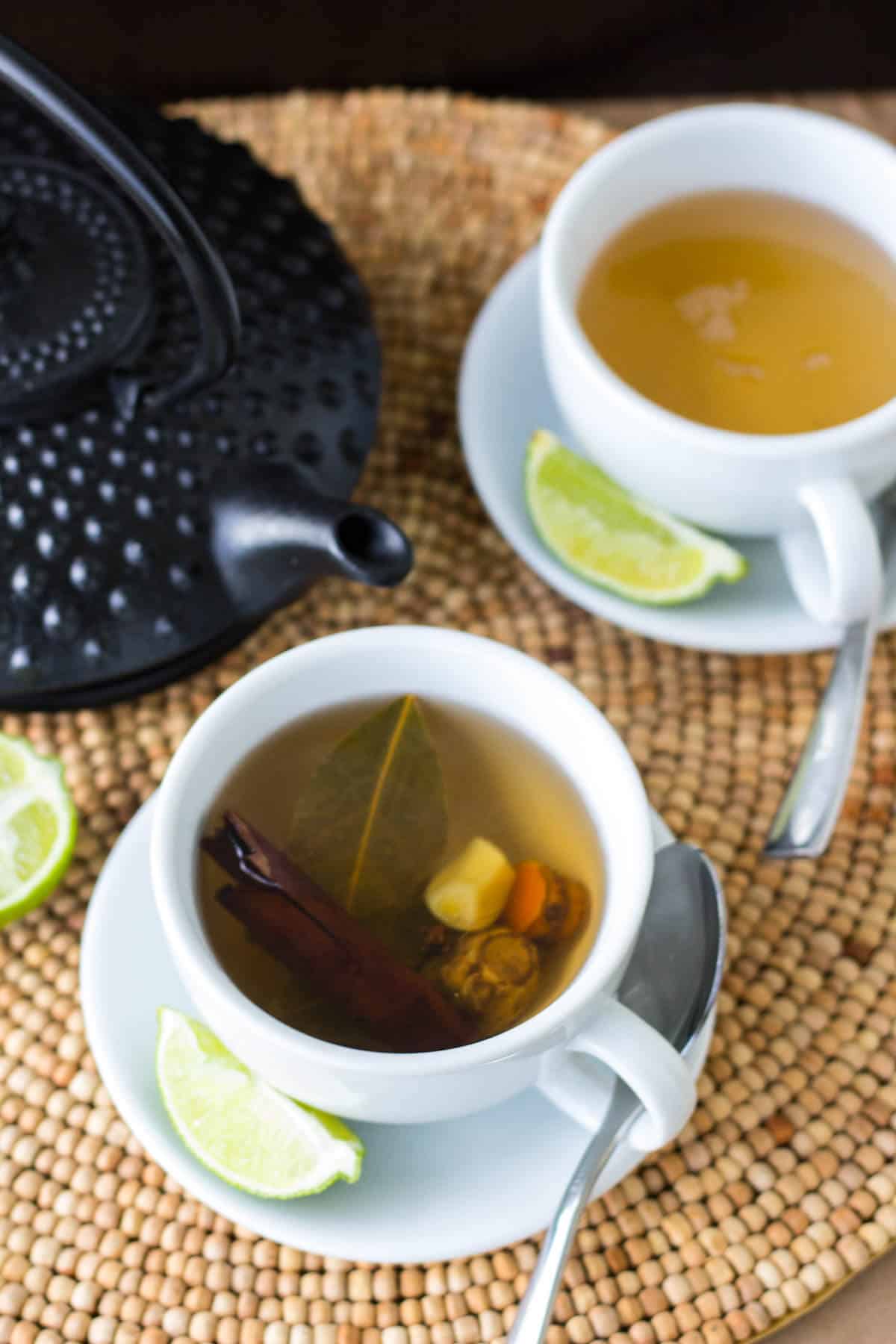 Two cups of homemade Caribbean bush tea served with fresh lime wedges and a black teapot.