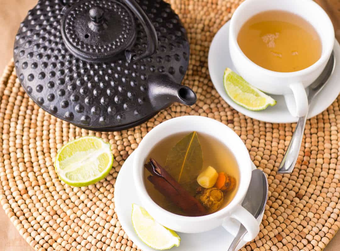 Caribbean bush tea served in white cups with a teapot and fresh lime wedges on a woven placemat.