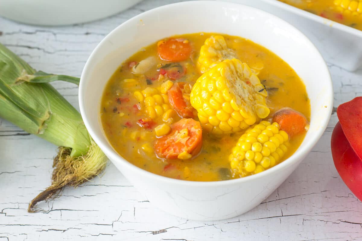 Trinidad corn soup is a popular Trini street food that is ram packed with flavor. from split peas, corn and dumplings. Perfect after fete snack. Bowl of Trinidad and Tobago corn soup with corn discs, carrots, and herbs, photographed beside fresh corn for a rustic Caribbean presentation.