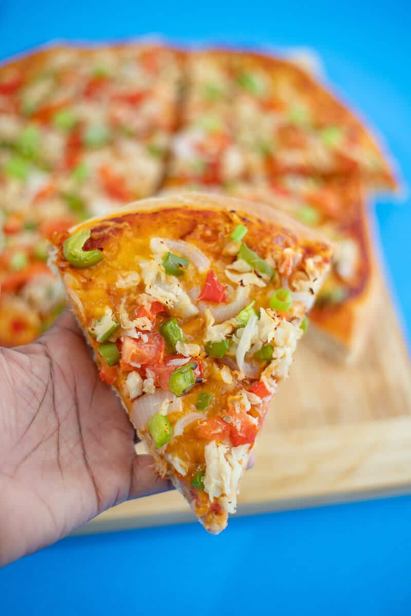 Hand holding a slice of homemade Caribbean saltfish pizza.