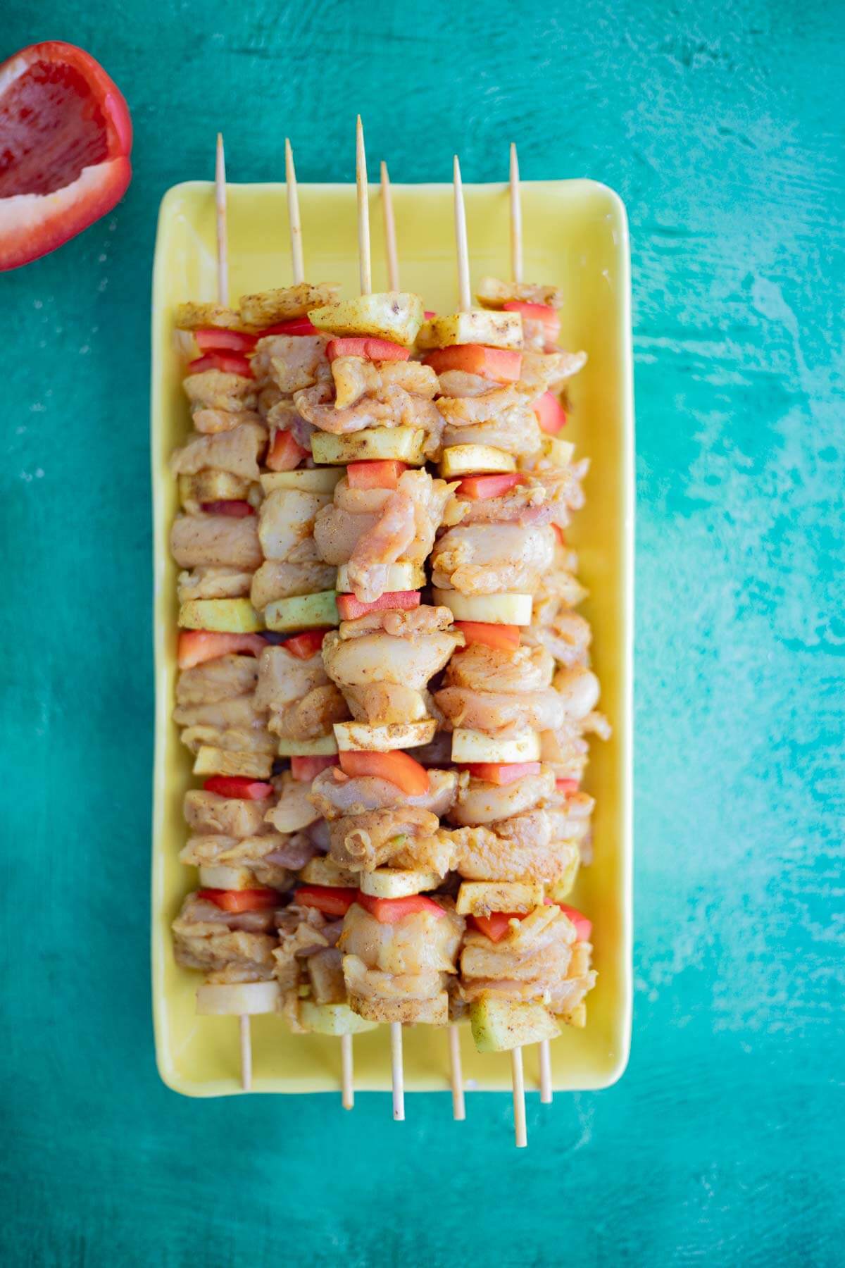 Five uncooked Caribbean chicken skewers with seasoned chicken, christophene, yellow squash, and red bell peppers arranged on a yellow tray.