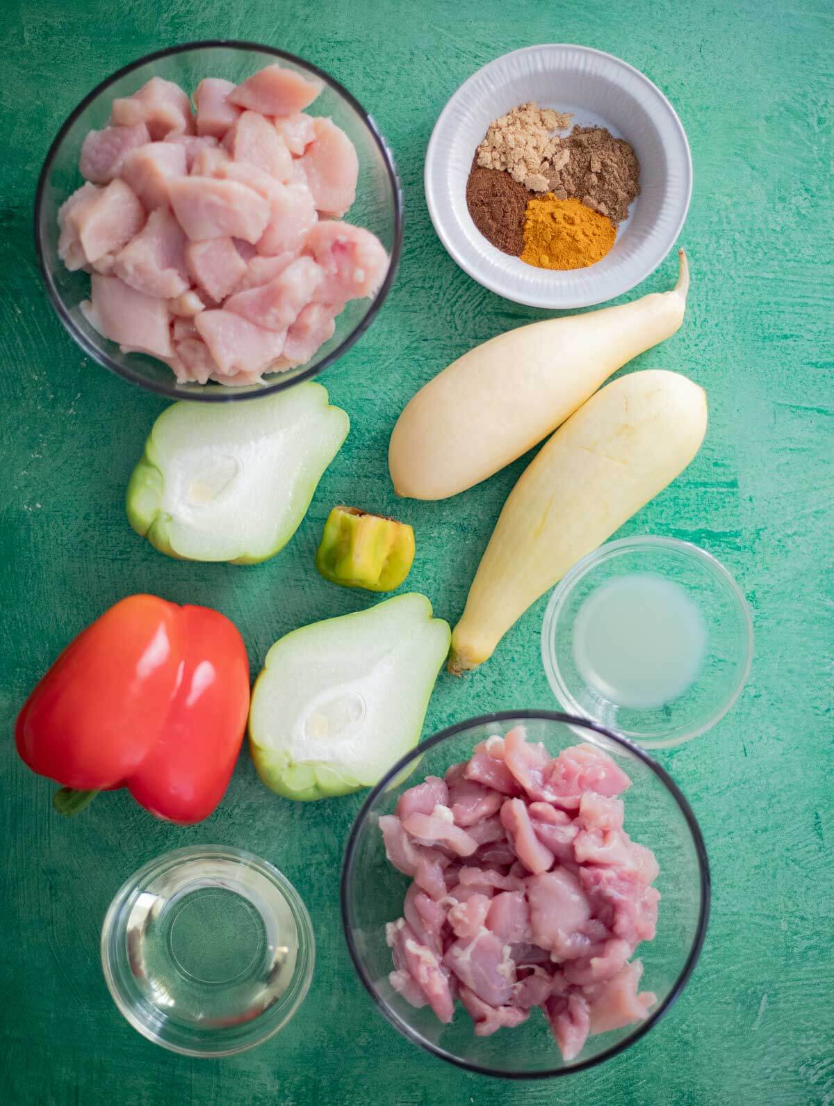 Raw chicken breasts and thighs, yellow squash, christophene, red bell pepper, spices, olive oil, and lime juice arranged on a turquoise background.