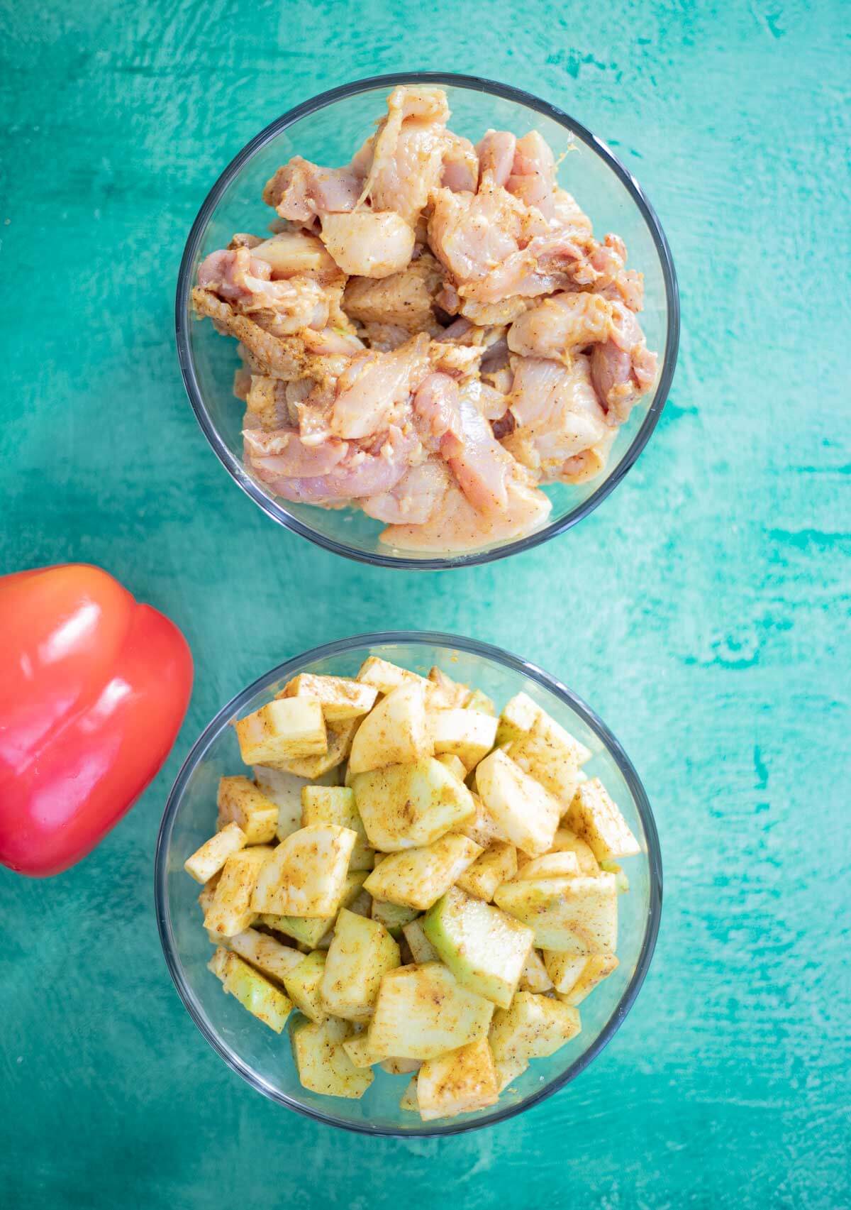 Two glass bowls containing raw chicken and chopped vegetables coated in a Caribbean spice marinade.