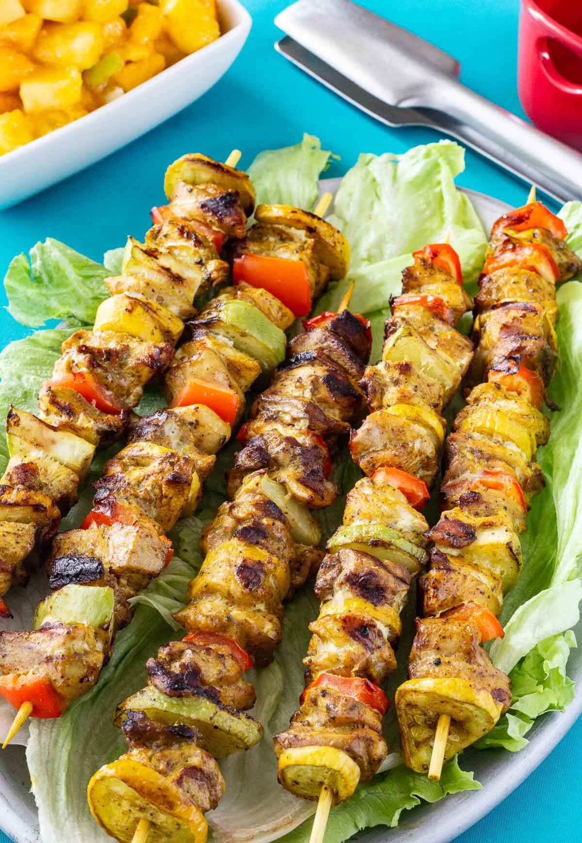 Grilled Caribbean chicken skewers with christophene, yellow squash, and red bell peppers arranged on a platter.