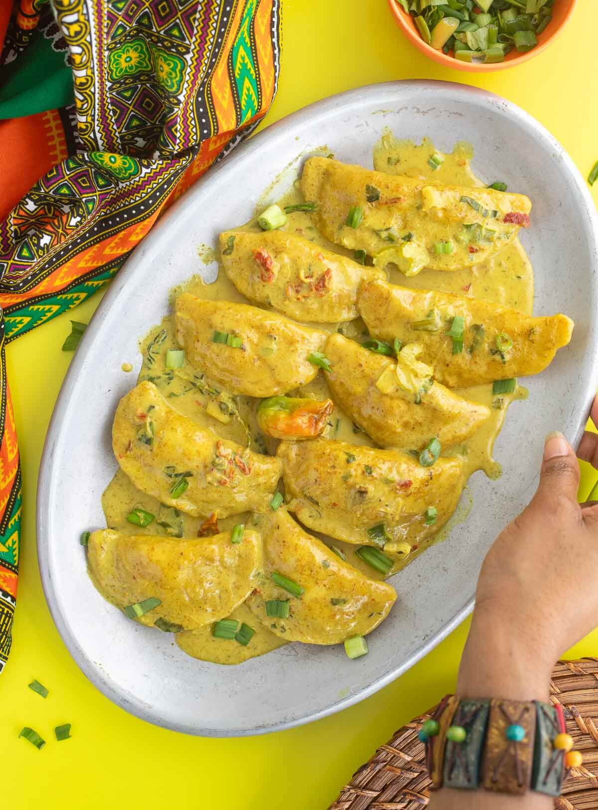 A upscale twist to the traditional dish, curry crab stuffed dumplings is the elevation you need. Dumplings stuffed with crab in a coconut curry sauce. Curry crab stuffed dumplings on a metal place with a african print cloth and a hand reaching for the plate. Additional scallion in a bowl for garnish.