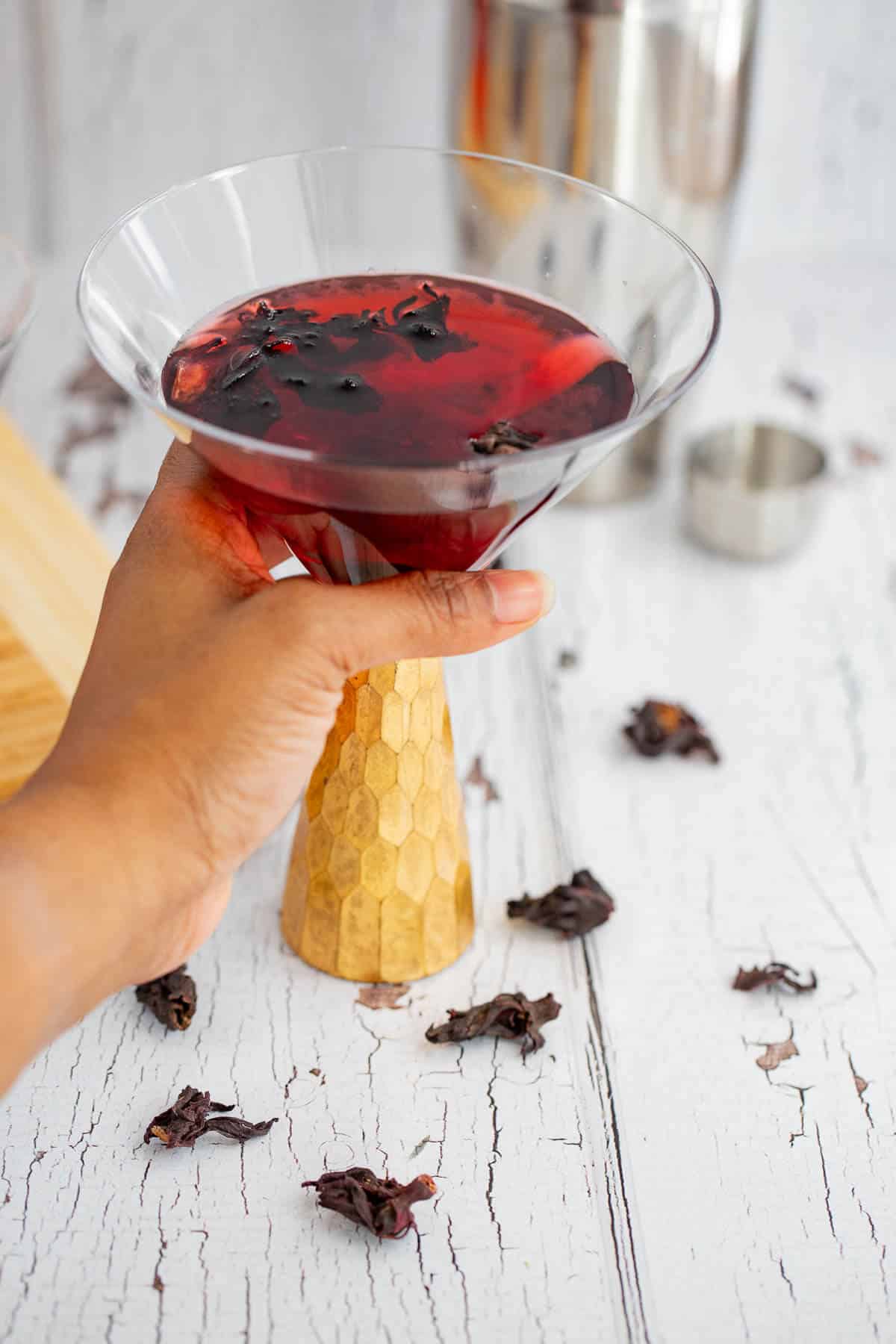 Hand holding a gold-stemmed glass filled with a deep red hibiscus martini.