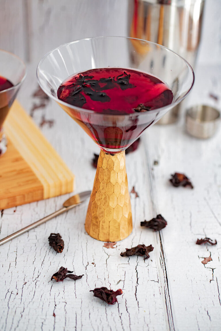 Picture of a martini glass with a gold handle filled with hibiscus martini with dried flowers sprinkled on the white background.
