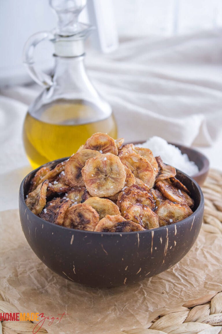 Plantain chips in a coconut bowl on a wooden table top with a bottle of oil in the background.