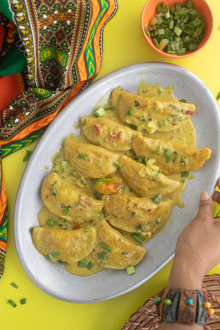 A picture of curry crab stuffed dumplings on a metal place with a african print cloth and a hand reaching for the plate. Additional scallion in a bowl for garnish.
