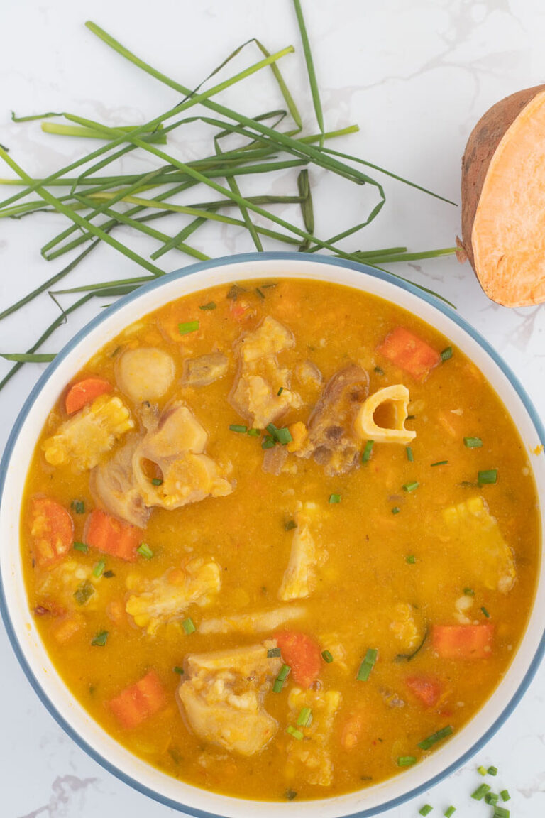 A blue bowl filled with Caribbean cow heel soup on a white counter top with some chives and a sweet potato in the background.