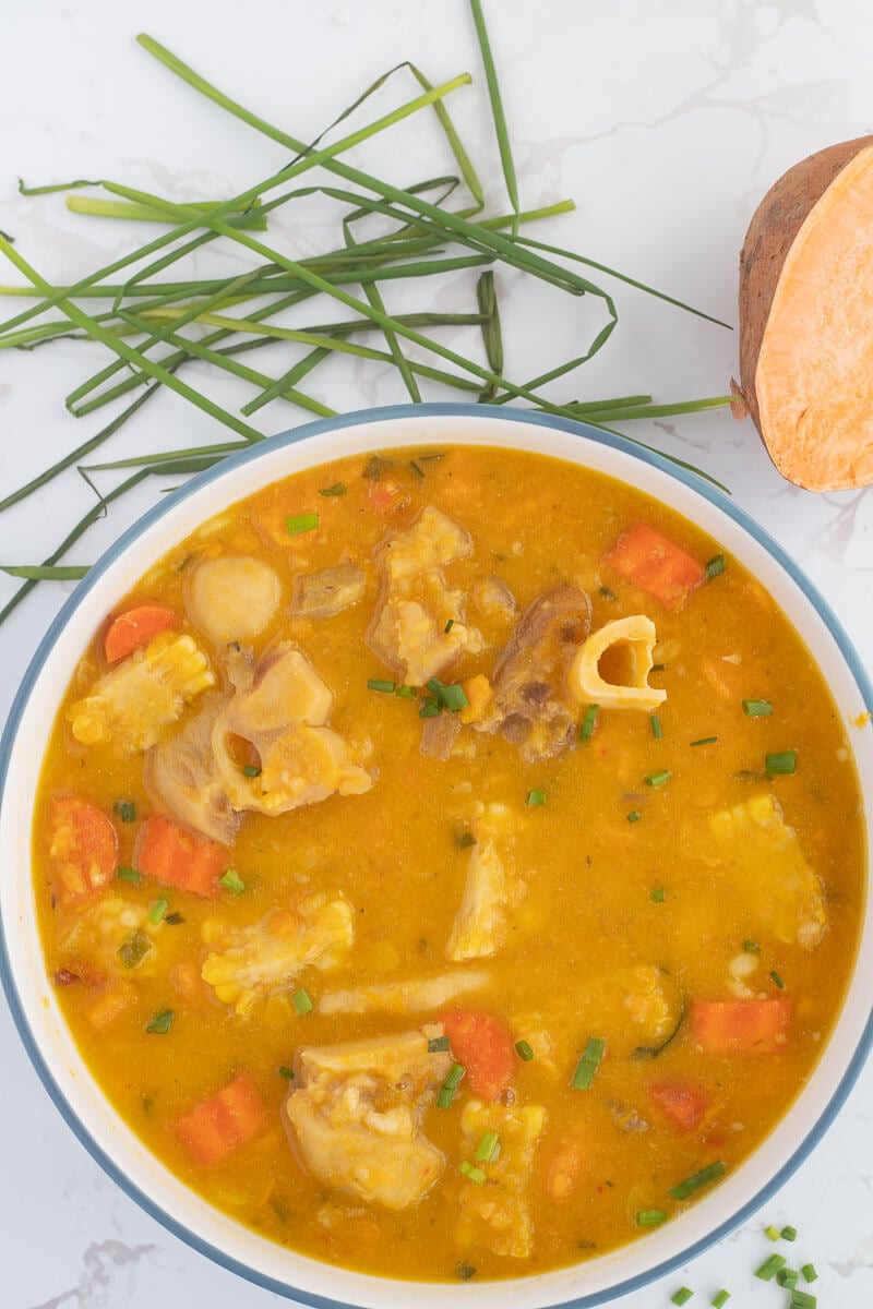 A blue bowl filled with Caribbean cow heel soup on a white counter top with some chives and a sweet potato in the background.
