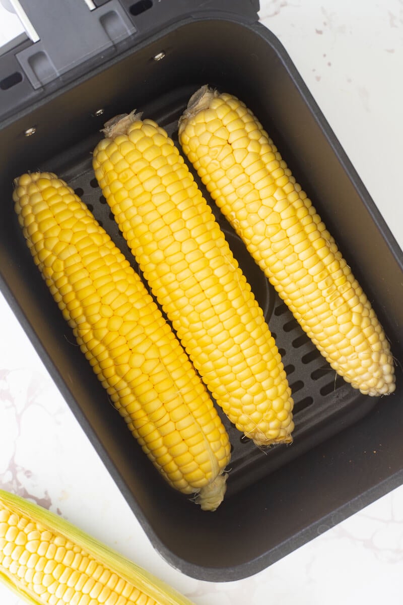 An airfryer basket with three cobs of corn in the basket.