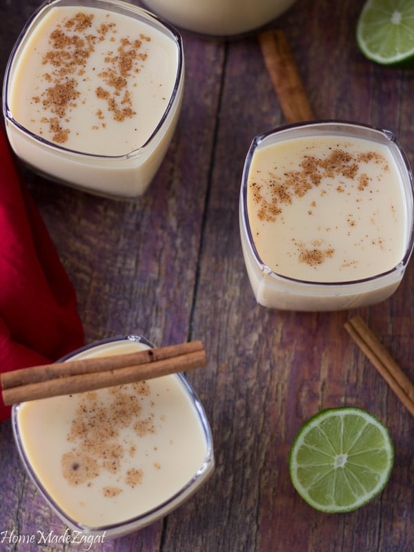 Glasses of ponche de creme on a brown background with a cinnamon stick and slice of lime.