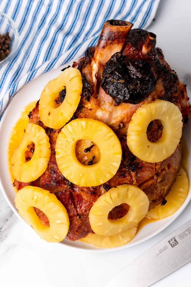 A leg of bone-in ham in a white plate, decorated with pineapples and cloves.