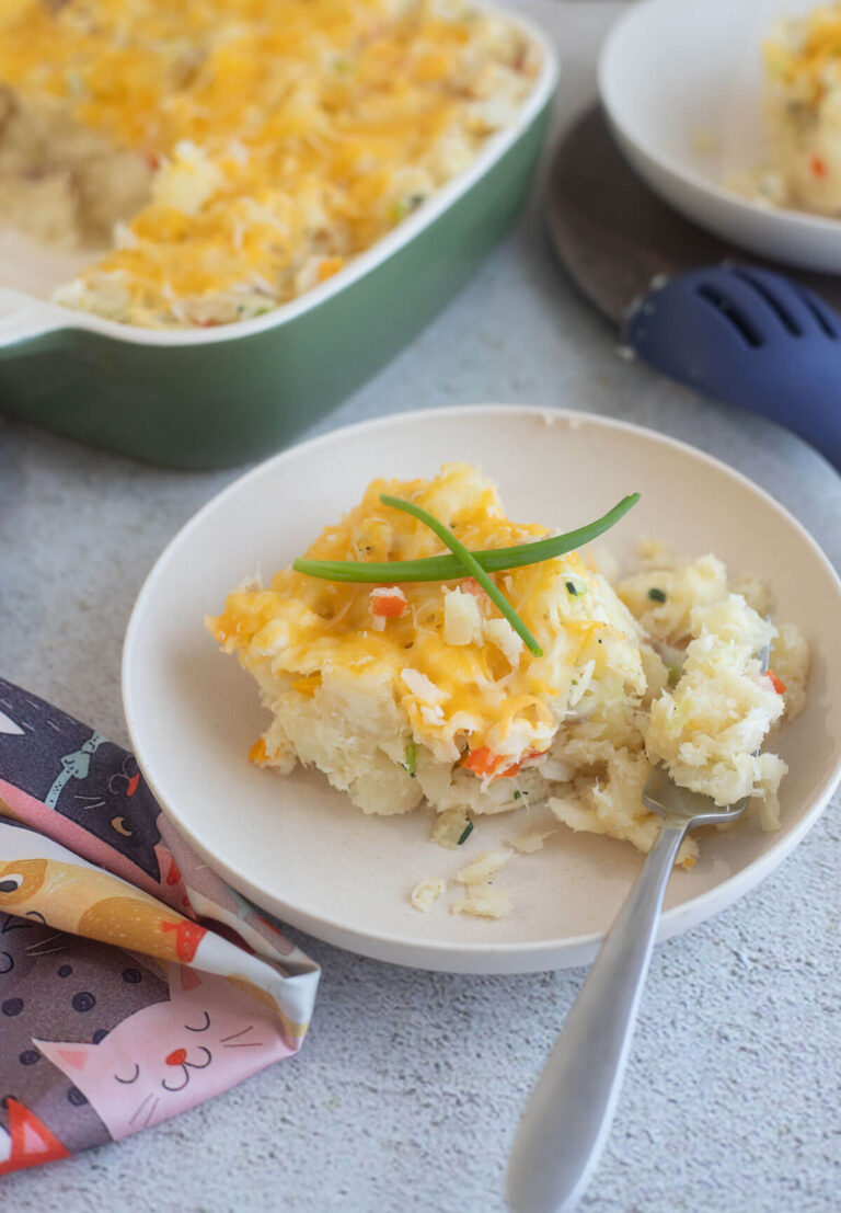 A slice of cassava and fish pie on a white bowl with a baking dish of fish casserole in the background.