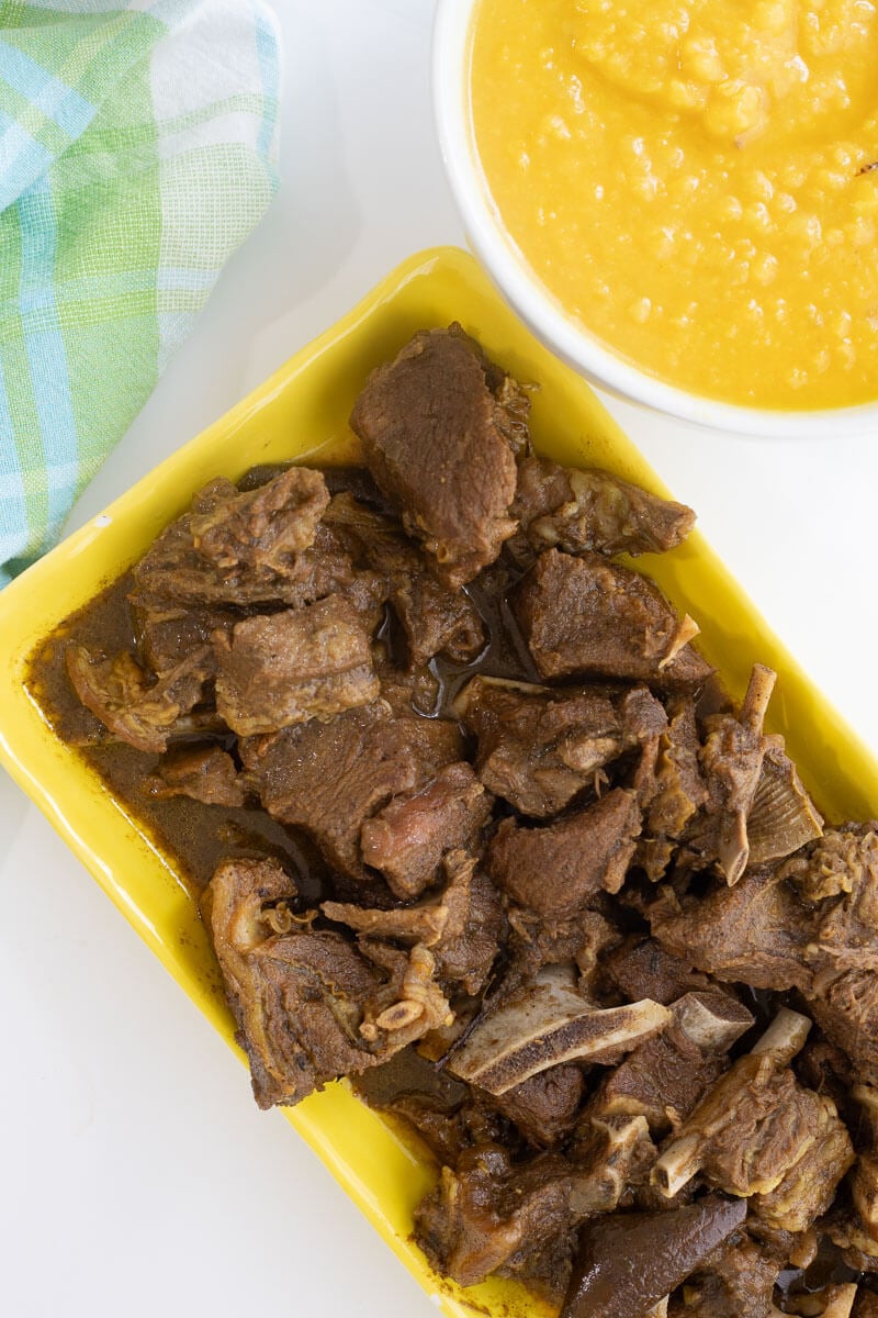 Cooked curry goat layed out on a yellow serving plate with a bowl of dhal and a patterned napkin on a white background.