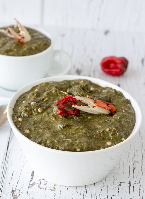 A large white bowl of crab and callaloo with a piece of crab set in the middle laying on a white counter top.