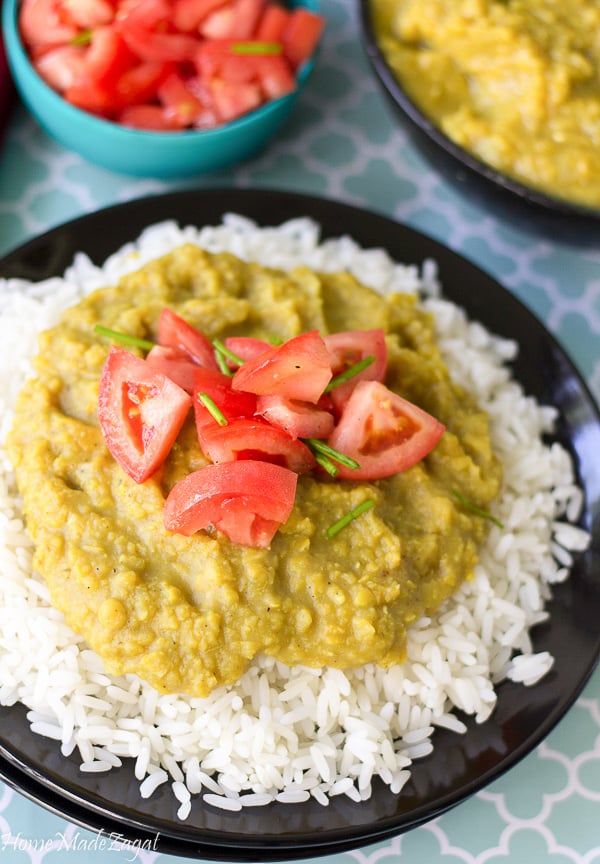 A chunky dhal with green split peas on top of white rice. And topped with chopped up peppers on a black plate. With more tomatoes in the background.