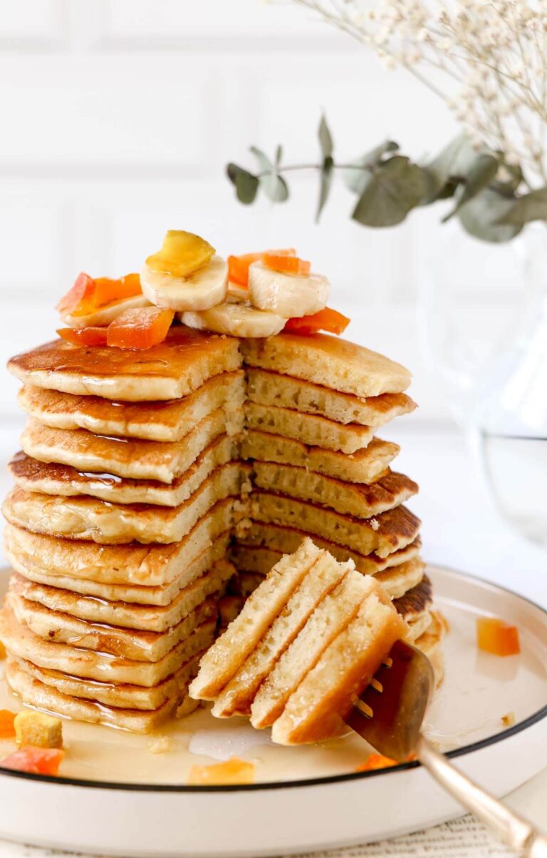 A high stack of pancakes on a white plate, topped with fruits and syrup.