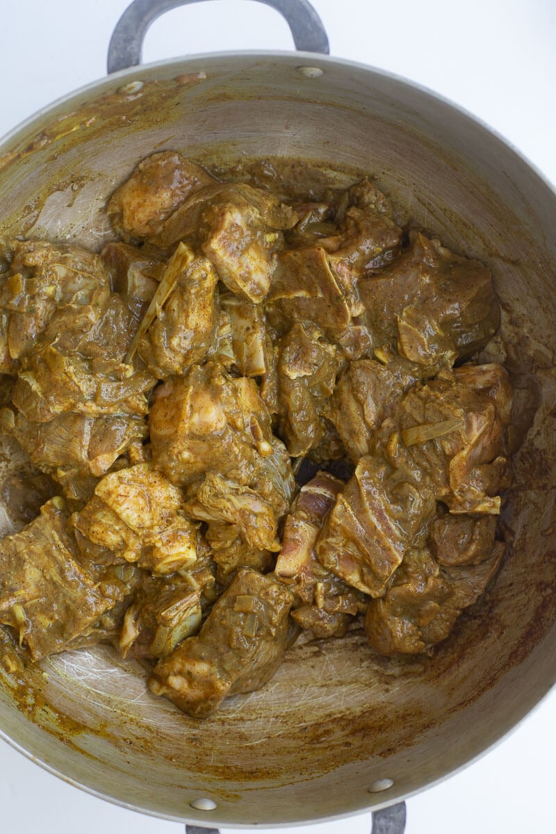 Seasoned goat meat added to a metal pot with aromatics.