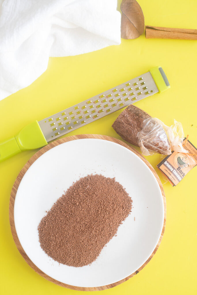 Overhead View of Grated Cocoa on a White Plate with a Grater and Cocoa Piece.