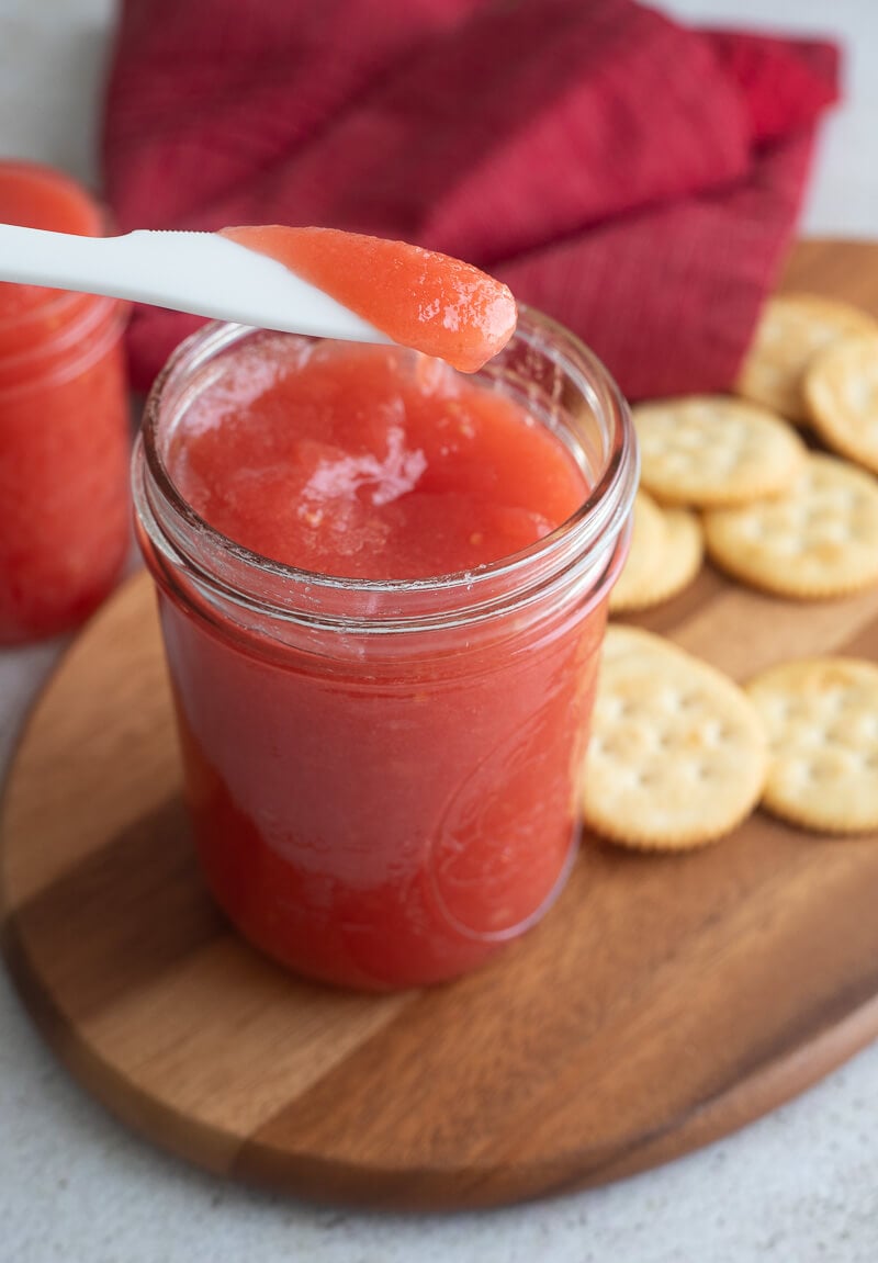 Guava jam on a knife hanging over a bottle of jam on a wooden bread board.