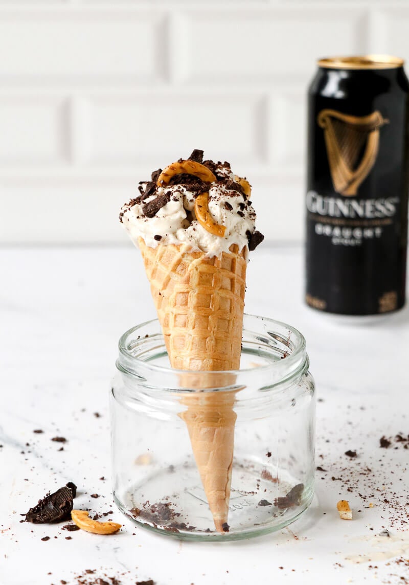 A cone of Guinness ice cream with sprinkles and pretzels with a can of Guinness in the background.
