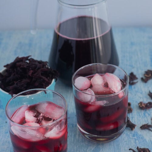 A jug of Caribbean sorrel drink with two glasses of sorrel with ice.