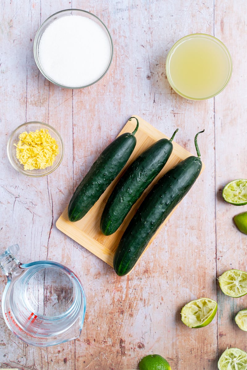 Fresh cucumbers, lime, ginger, and a jar of water on a countertop, ready to make homemade cucumber and lime juice.
