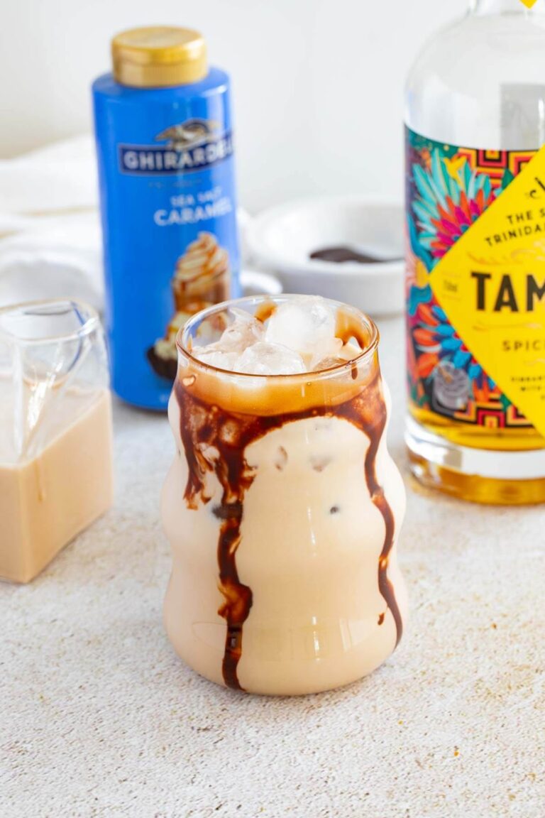A glass filled with Jamaican rum creme, lined with chocolate with a blue bottle in the background of caramel sauce and a smaller bottle with rum creme.