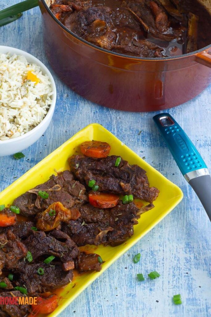 Hearty lamb stew slices on a yellow plate with a pot of servings and a bowl of rice and okra