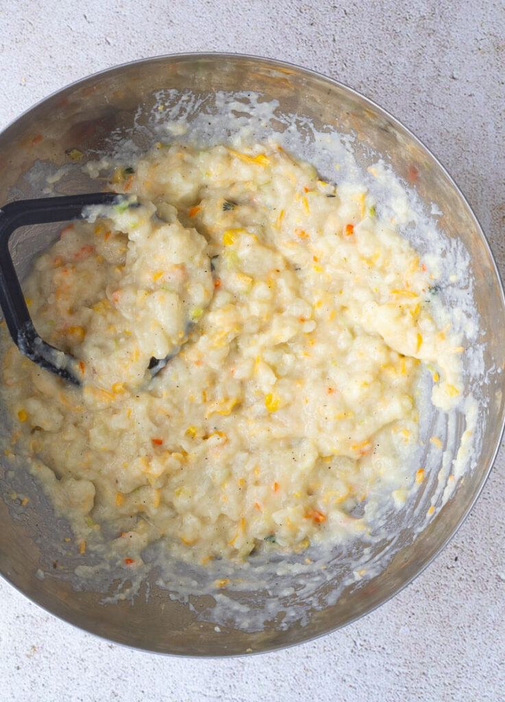 A bowl of mashed potatoes with creamed corn and a black potato masher.