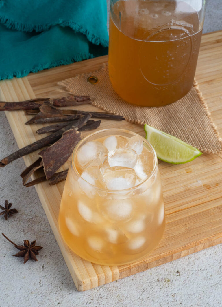 Mixed mauby drink in a glass of pebbled ice with star anise and mauby bark in the background.