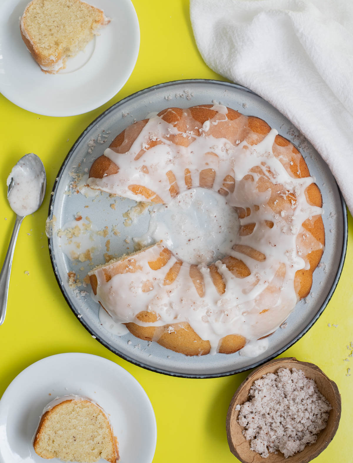 A coconut cake with slices of cake on white plates with shredded coconut and spoons of glaze