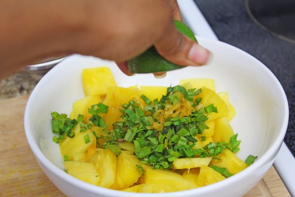 Lime juice being squeezed over a white bowl of chopped up pineapples with fresh herbs.