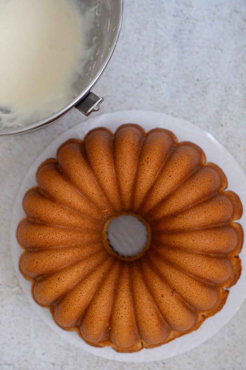 Bundt cake on a cake board with a silver bowl of ponche de creme glaze.