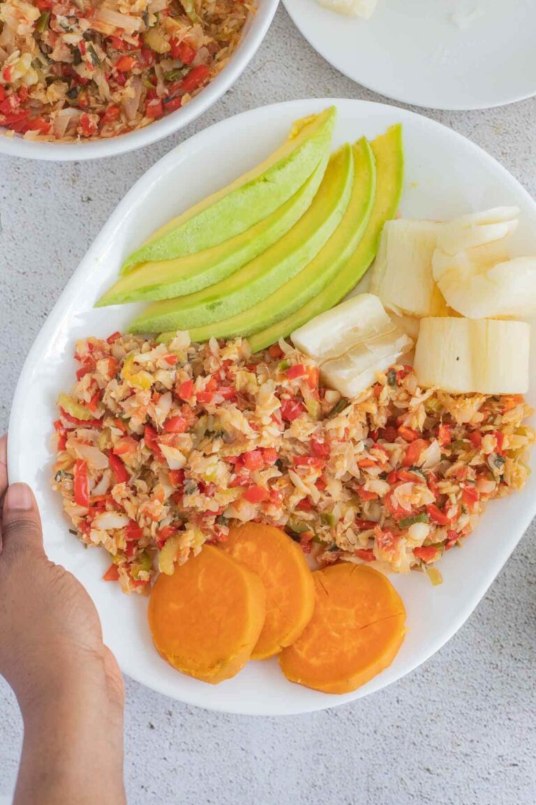 A plate of stew saltfish with ground provision on a white plate. And a serving bowl of stewed saltfish on the side.