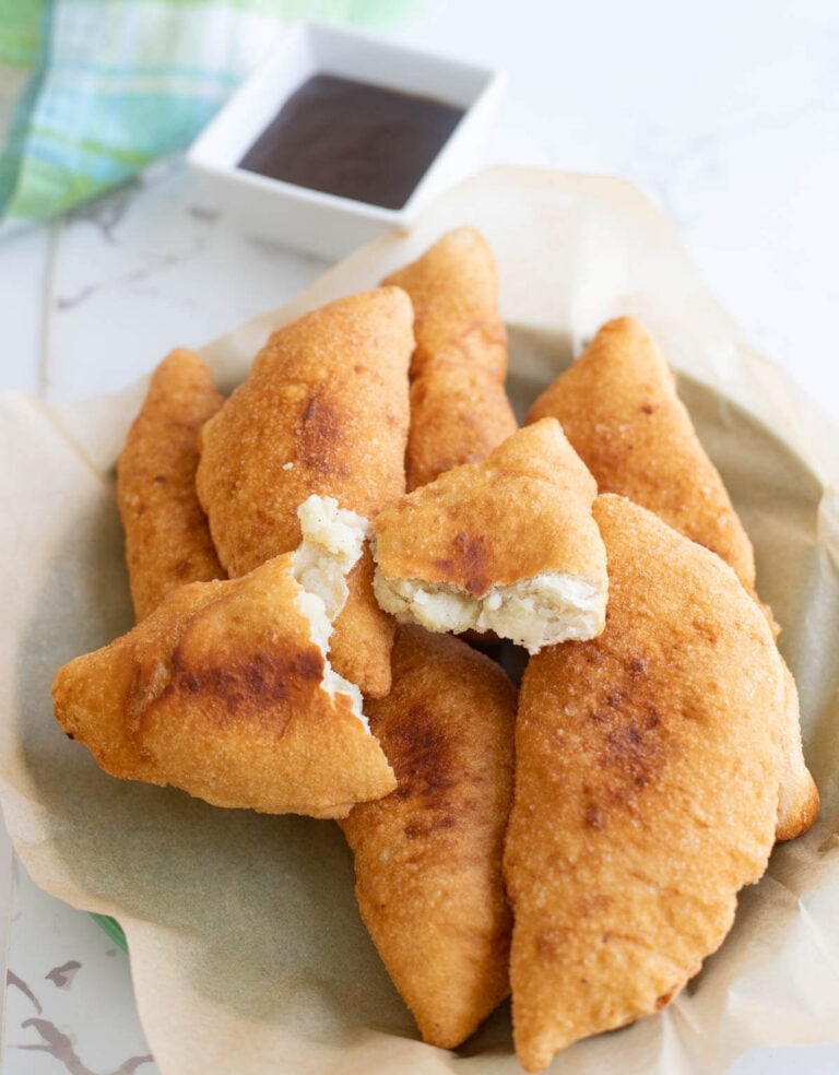 A bunch of fried aloo pies on a plate with wax paper on a white background with tamarind sauce in a bowl and a checkered napkin in the background.