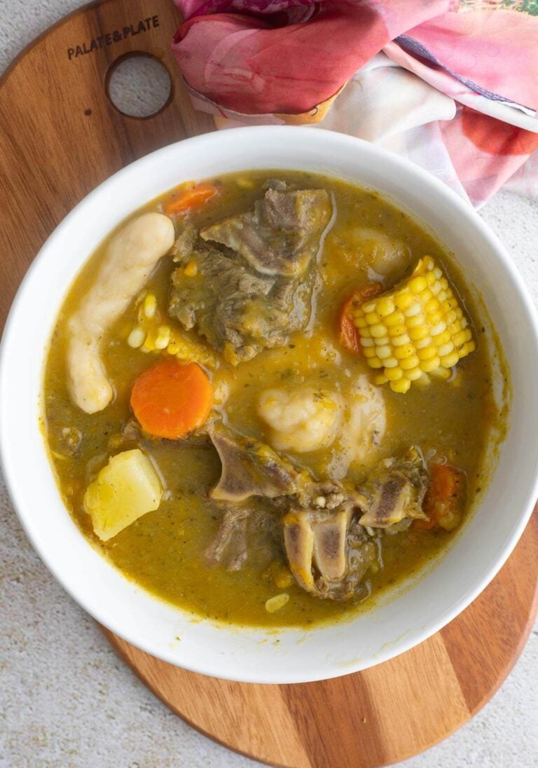 A white bowl of soup with beef, dumplings and corn on a wooden board with a patterned cloth.
