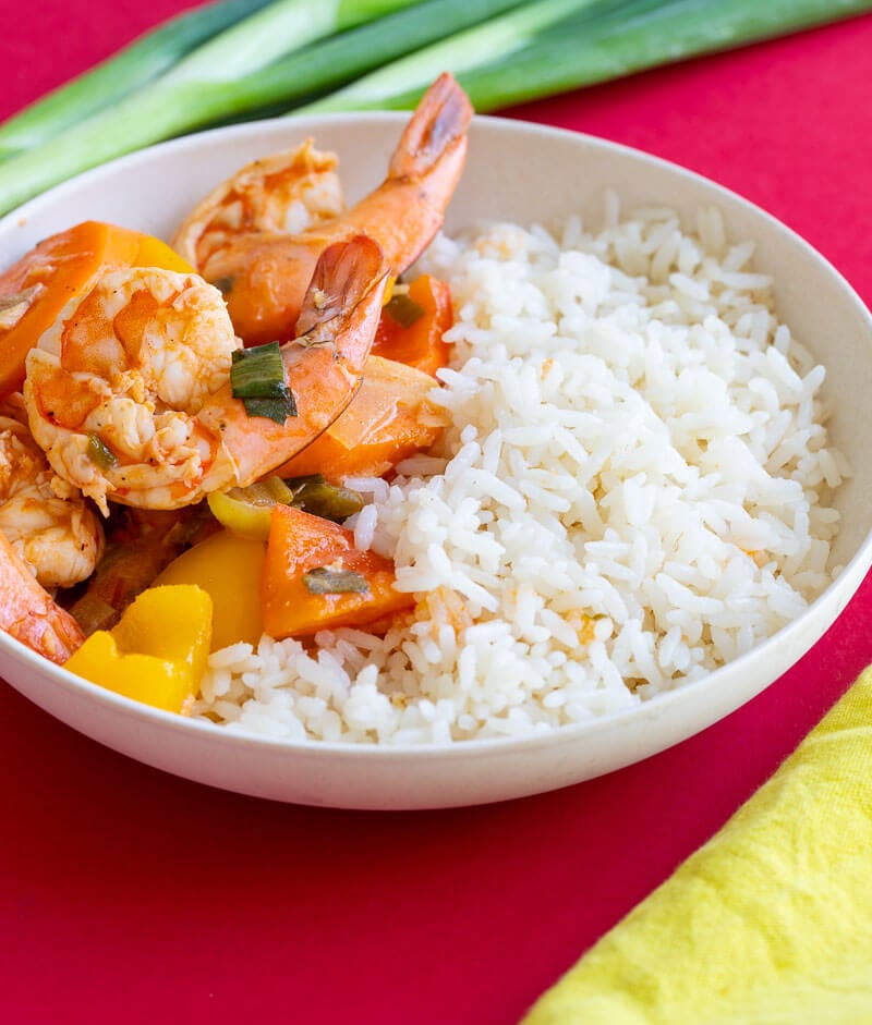 A bowl of pepper shrimp and rice on top a red counter with a yellow napkin.
