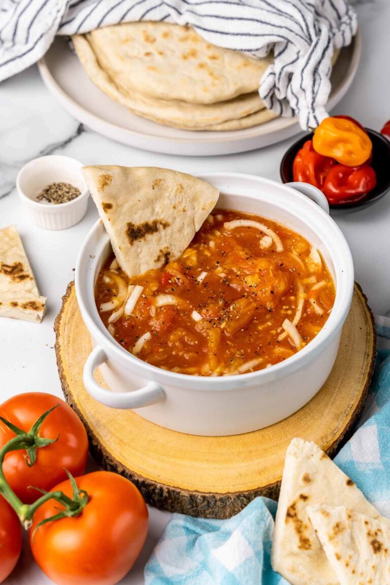 A white pot of trinidad tomato choka with sada roti dipped in the bowl and salt and pepper on the side to season.