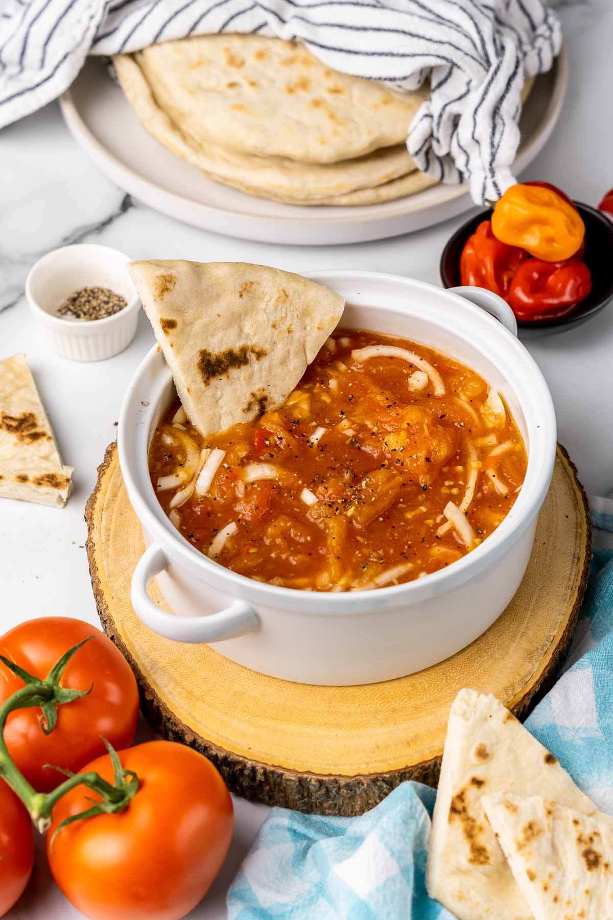 A white pot of trinidad tomato choka with sada roti dipped in the bowl and salt and pepper on the side to season.