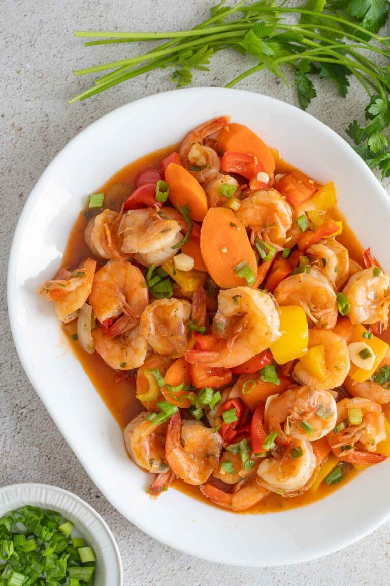 An overhead view of a white plate filled with pepper shrimp and bowl of chopped scallions on the side to garnish.